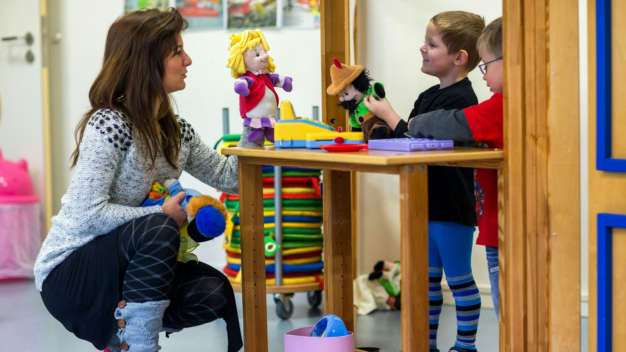 Eine Frau und zwei Kinder spielen mit Handpuppen. Die Frau hockt auf dem Boden vor den Kindern.