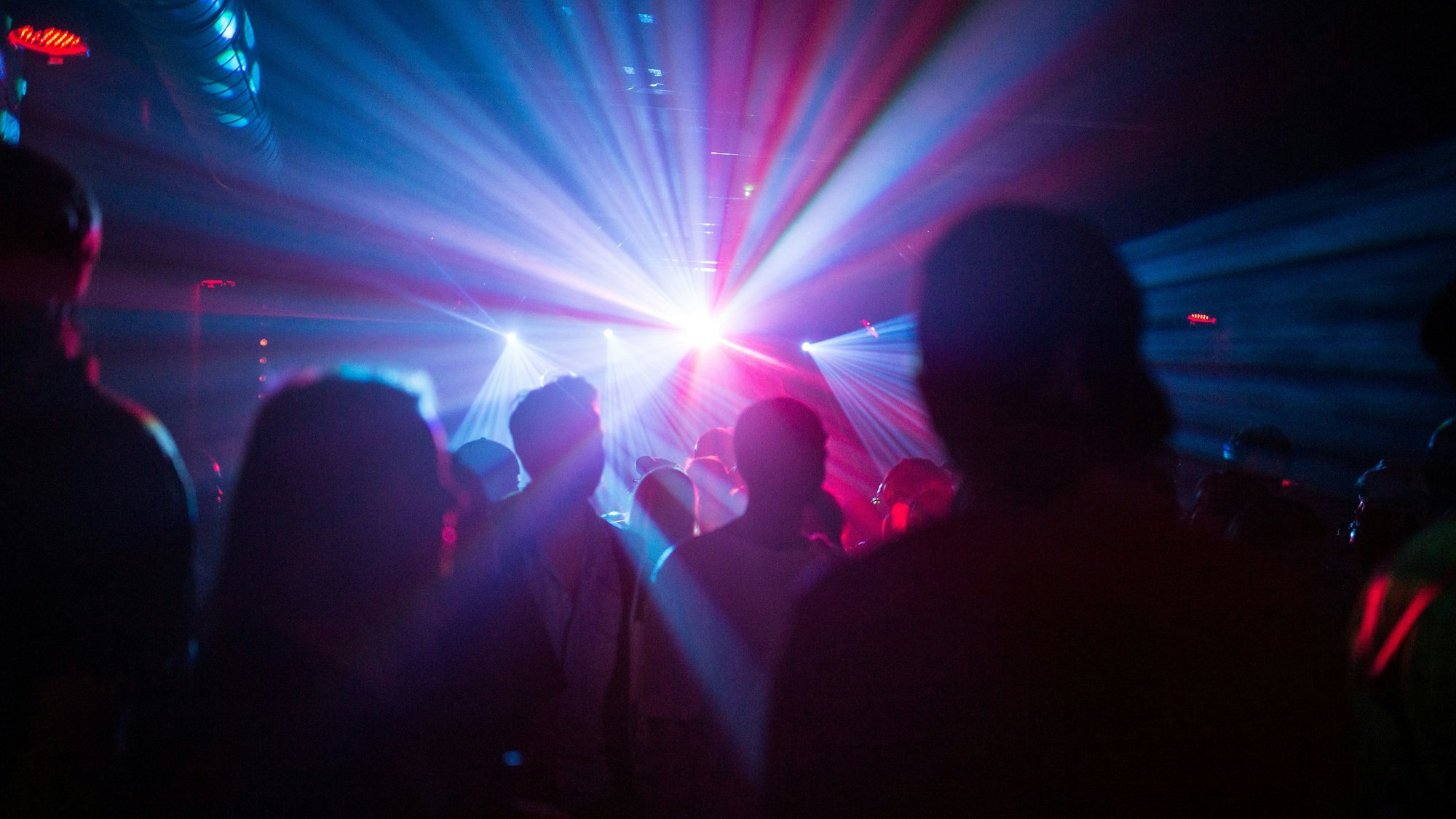 Eine Gruppe von Menschen steht mit dem Rücken zur Kamera in einem Club und wird von Lichtstrahlern angeleuchtet.
