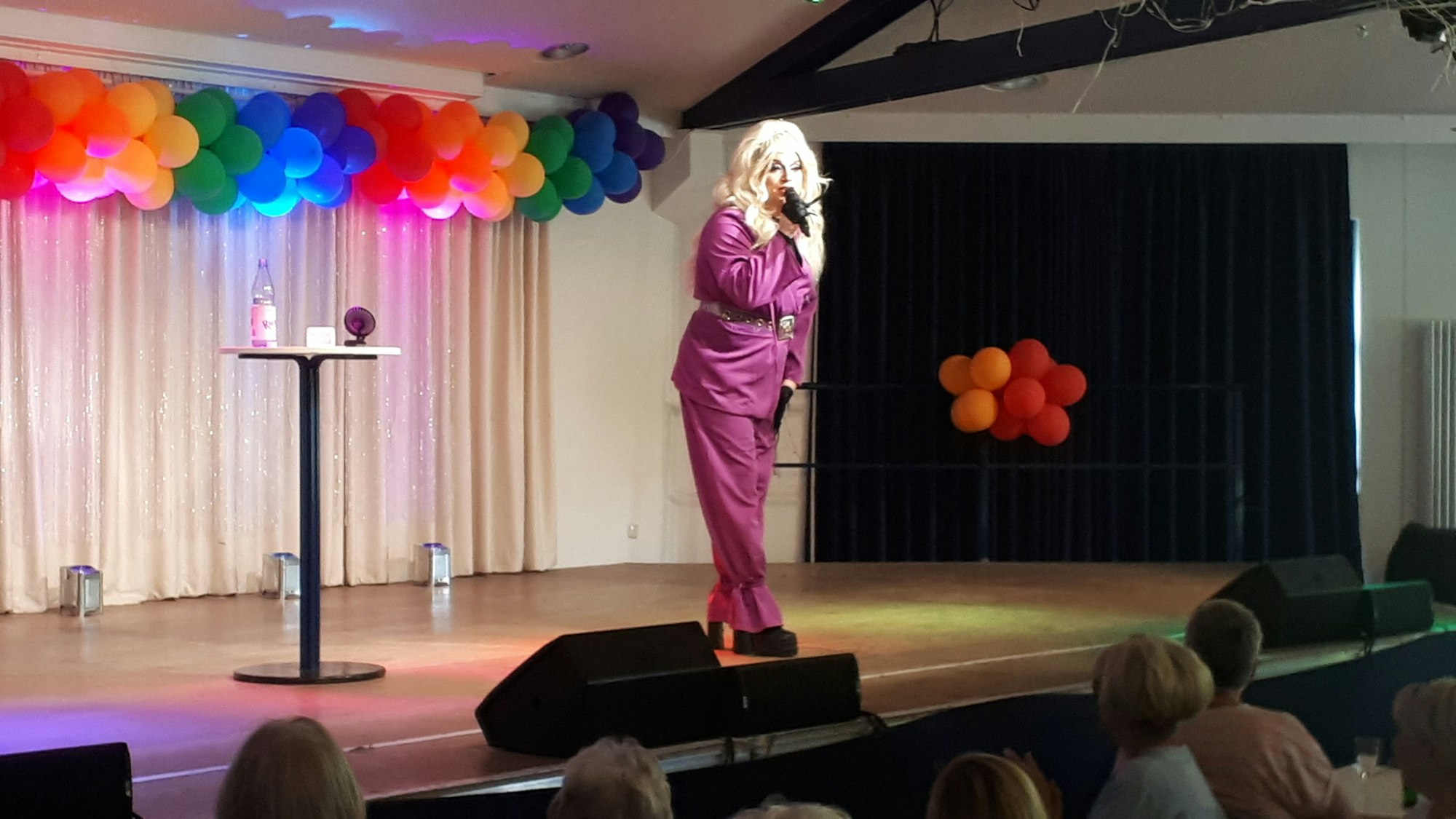 Eine Dragqueen mit langen blonden Haaren im lilafarbenen Anzug steht auf einer Bühne. Im Hintergrund ist ein Vorhang zu sehen, daran befestigt sind regenbogenfarbene Ballons.
