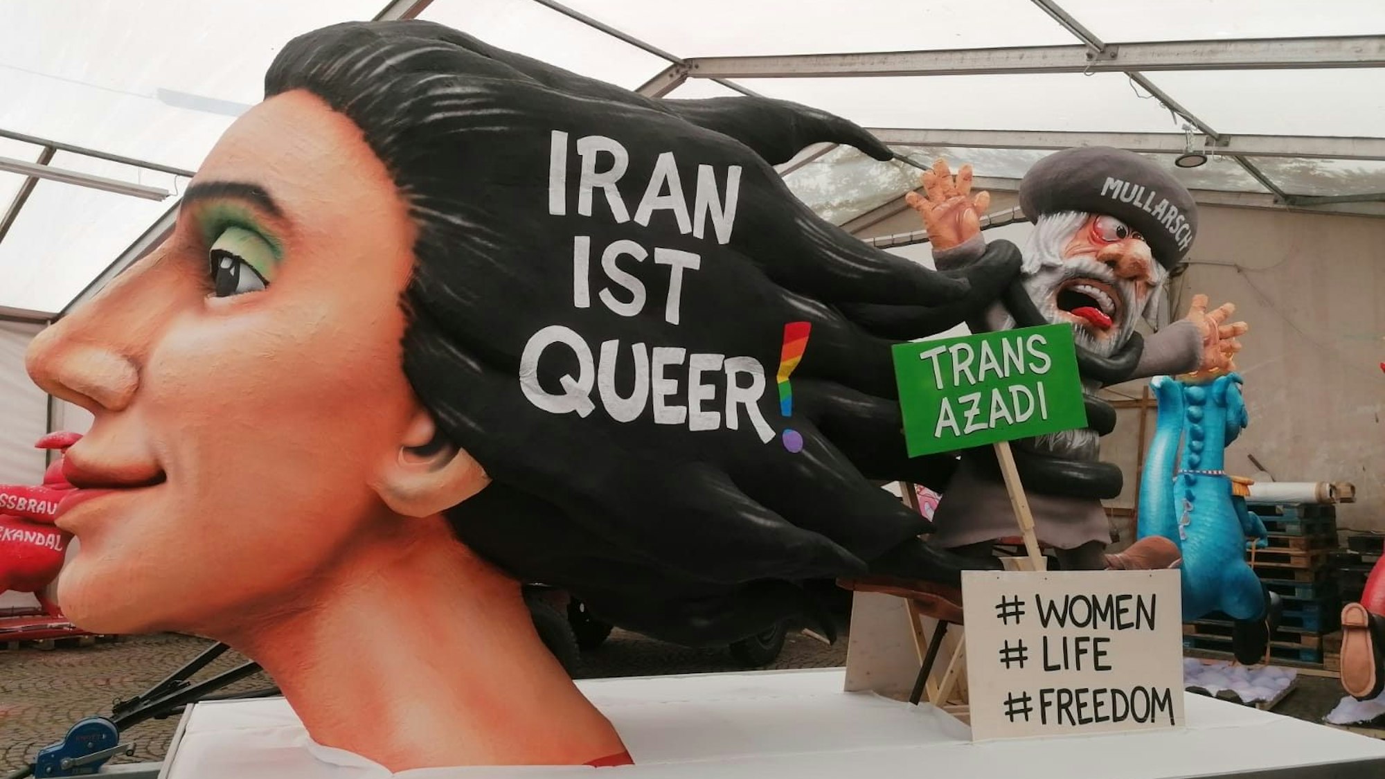 Der Woman-Life-Freedom-Wagen auf dem Kölner CSD wurde von Jaques Tilly gebaut. Vorne wird ein Kunstwerk einer Frau sein, die mit ihren Haaren einen Mullah erwürgt. Zusätzlich steht "Iran ist queer!" in ihren Haaren. Auf einem grünen Schild steht außerdme „Trans Azadi“, auf einem weiteren „#Women #Life #Freedom“.