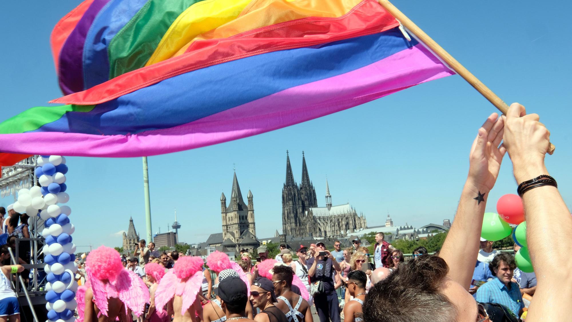 Jemand schwingt eine Regenbogenflagge. Im Hintergrund ist der Kölner Dom zusehen.
