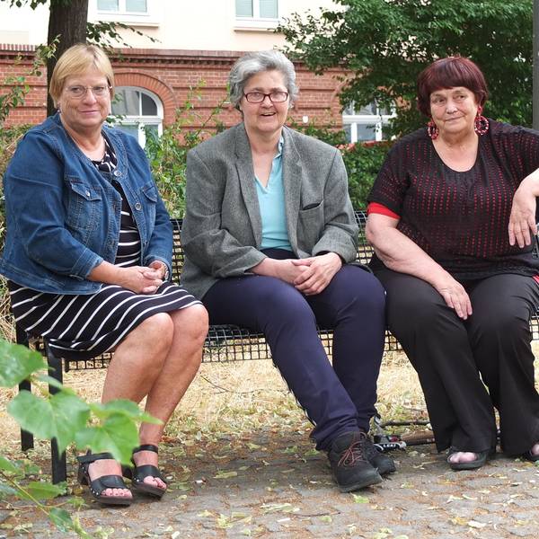 Drei ältere Damen sitzen auf einer Bank, eine stützt den Arm auf ihren Rollator.