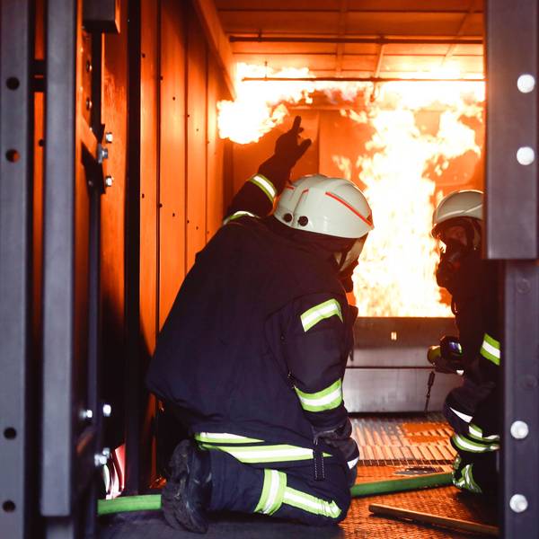 Zwei Männer in Feuerwehruniformen knien im Brandcontainer der Feuerwehr, im Hintergrund sind Flammen zu sehen.