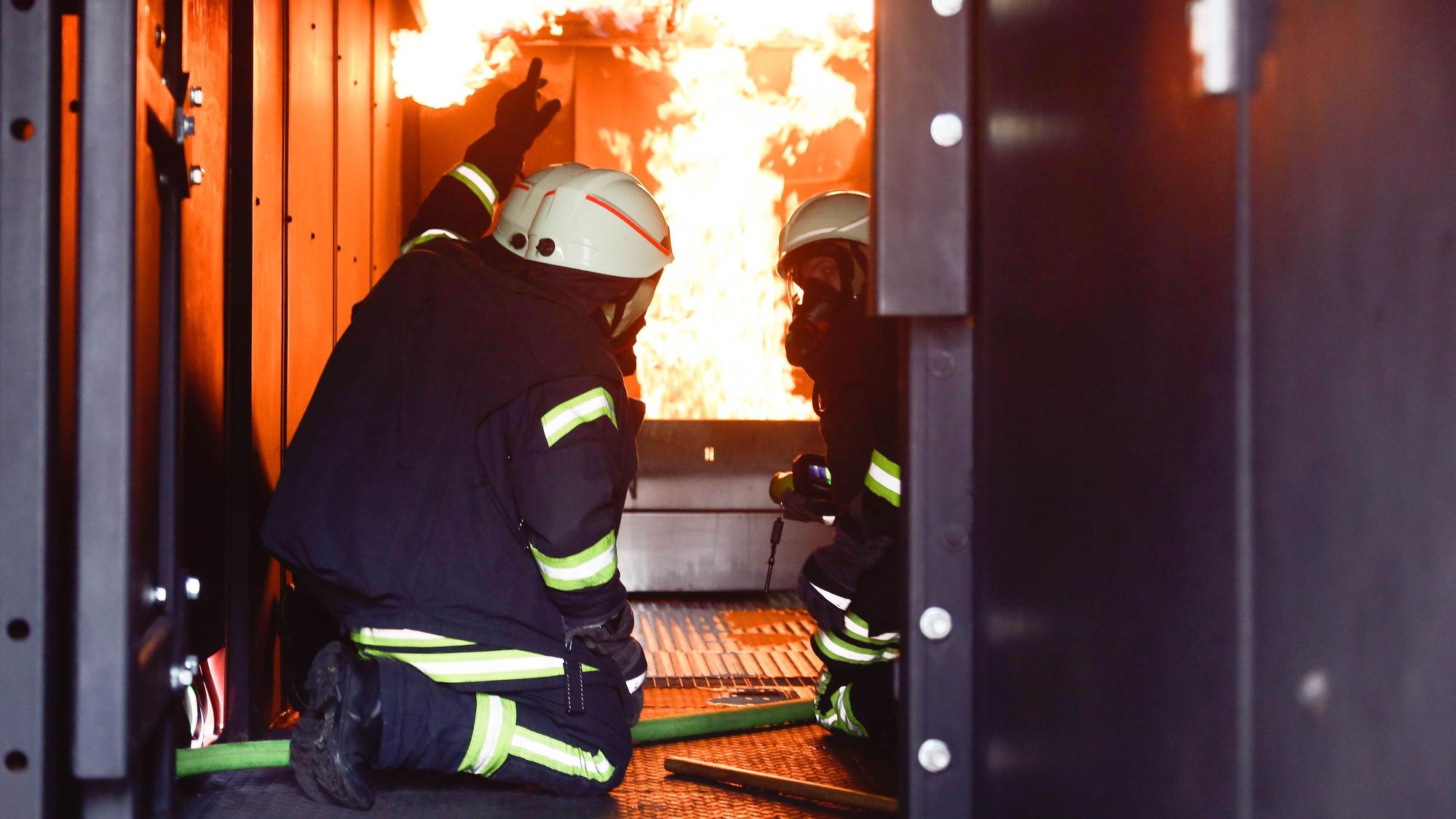 Zwei Männer in Feuerwehruniformen knien im Brandcontainer der Feuerwehr, im Hintergrund sind Flammen zu sehen.