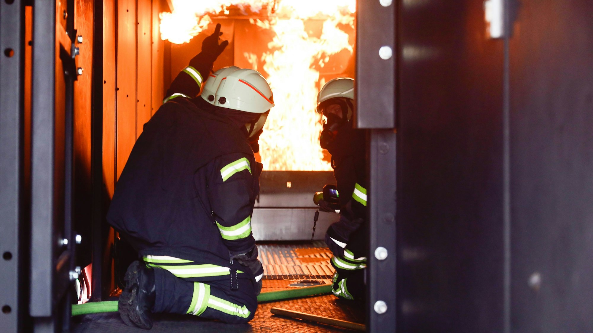 Zwei Männer in Feuerwehruniformen knien im Brandcontainer der Feuerwehr, im Hintergrund sind Flammen zu sehen.