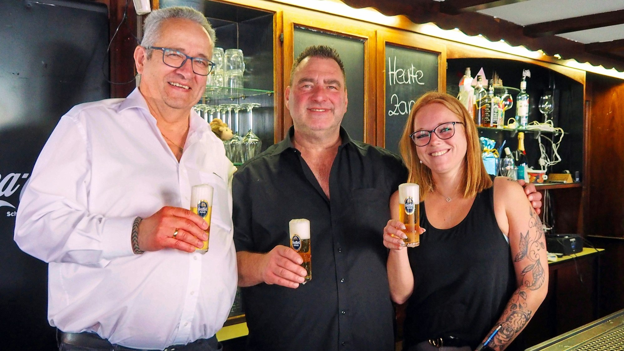 Zwei Männer und eine junge Frau stehen mit Kölsch in der Hand hinter einer Theke.