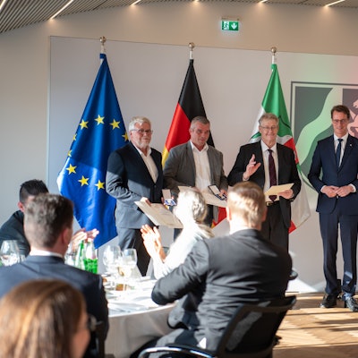 Wolf-Christian Lorenz, Carsten Bönsch, Hubert Schilles und Ministerpräsident Hendrik Wüst stehen in der Staatskanzlei vor einem den Flaggen von der Europäischen Union, Deutschland und Nordrhein-Westfalen. Sie werden von einem sitzenden Publikum beklatscht.