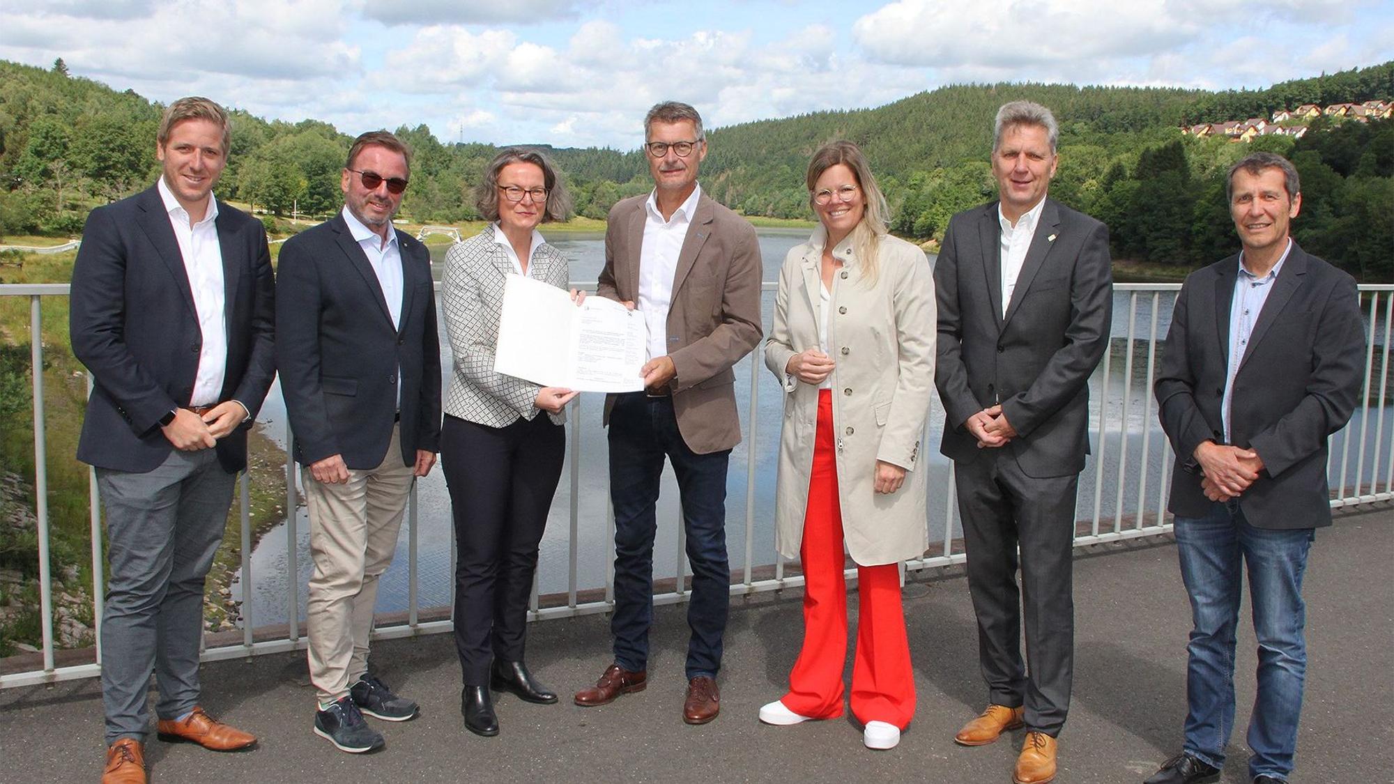 Die Ministerin überreicht Bürgermeister Lembach auf dem Damm des Kronenburger Sees den Bescheid, daneben die Landräte und kommunale Vertreter.