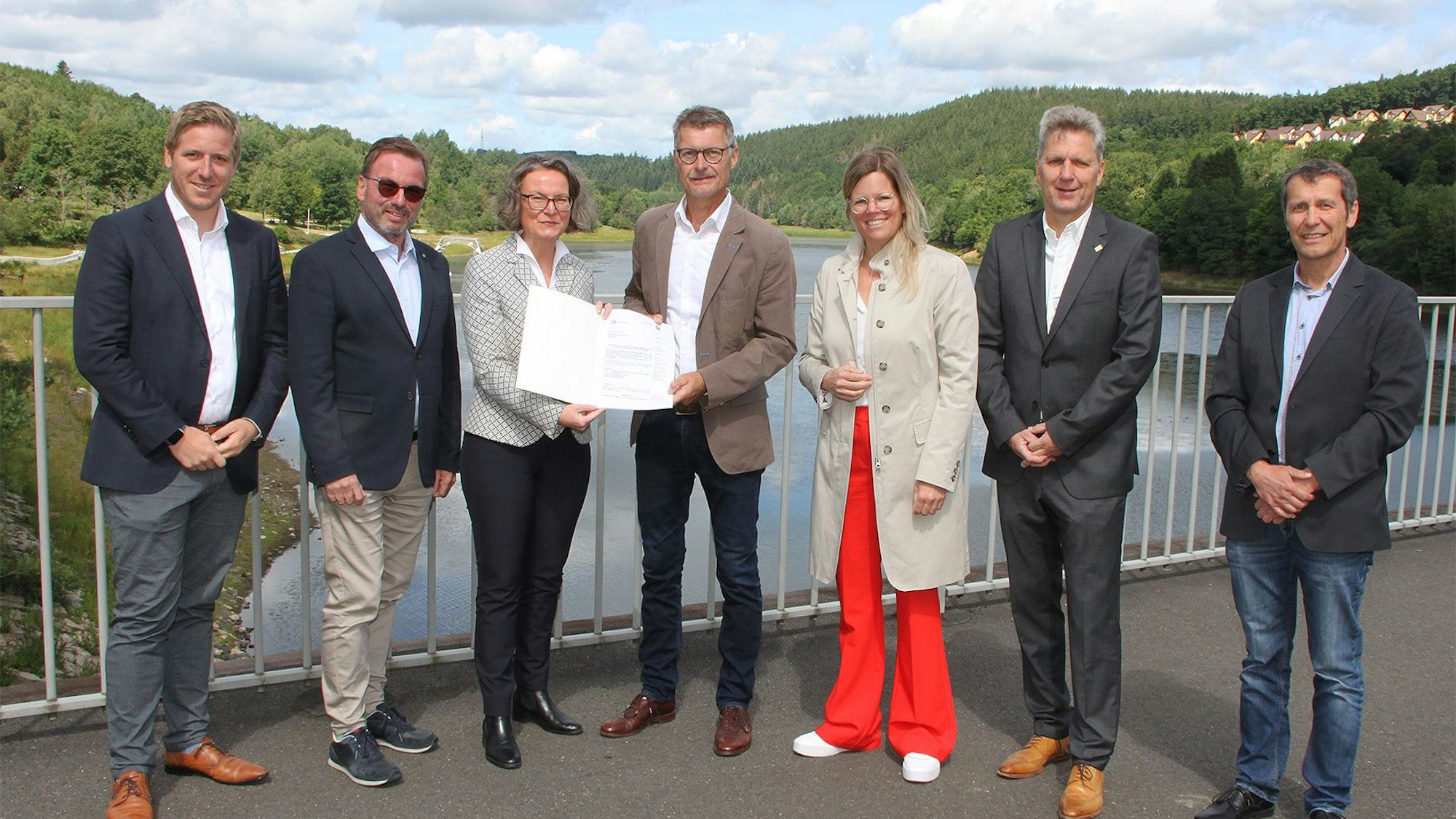 Die Ministerin überreicht Bürgermeister Lembach auf dem Damm des Kronenburger Sees den Bescheid, daneben die Landräte und kommunale Vertreter.