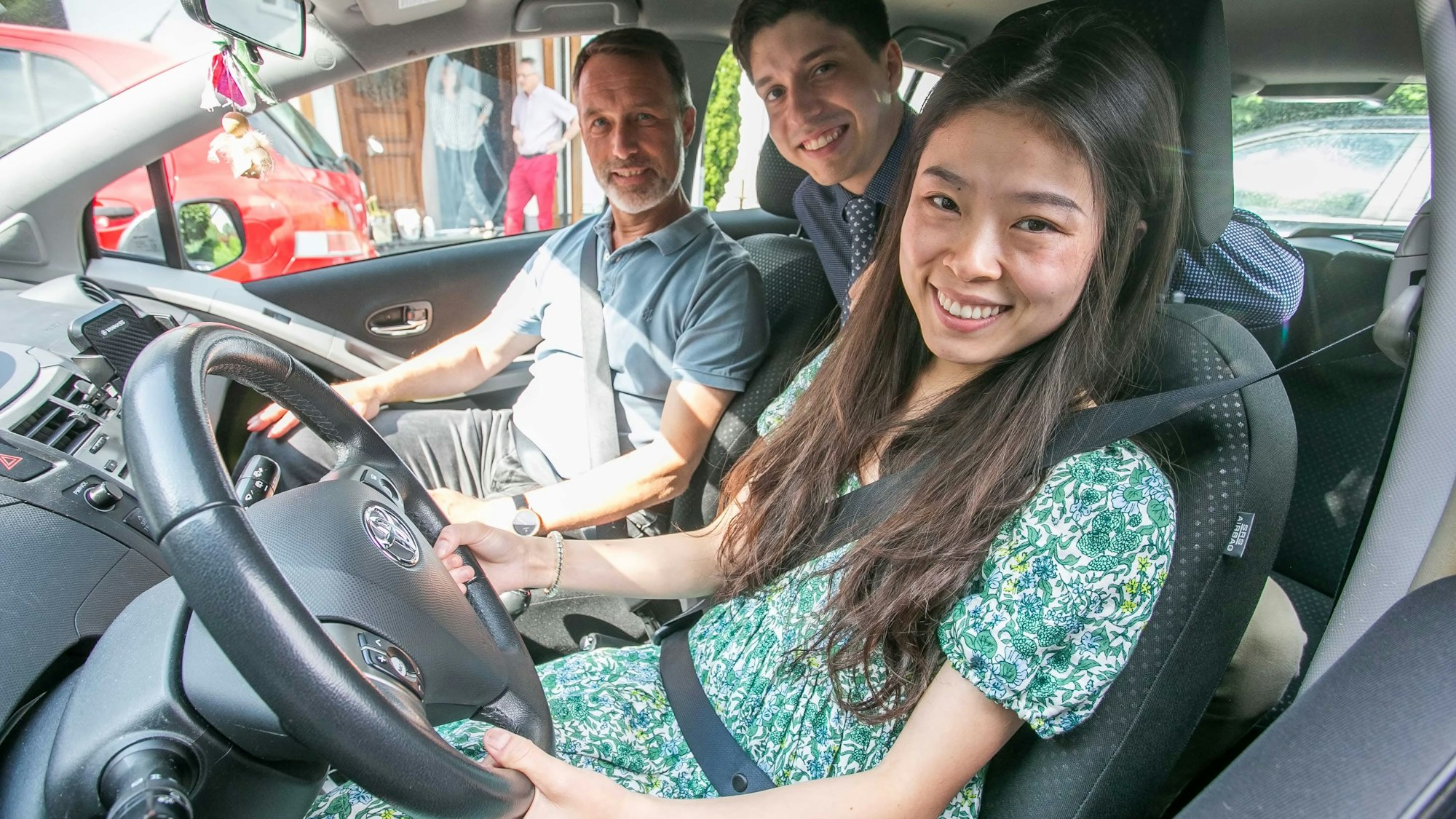 Die Pianistin Mimi Zhang sitzt hinter dem Lenkrad eines Kleinwagens mit Gangschaltung, auf der Rückbank ihr Lebensgefährte David Mach, auf dem Beifahrersitz Bernd Wegerhoff, den den beiden Meisterkurs-Schülern das Fahren mit Gangschaltung erklärt.