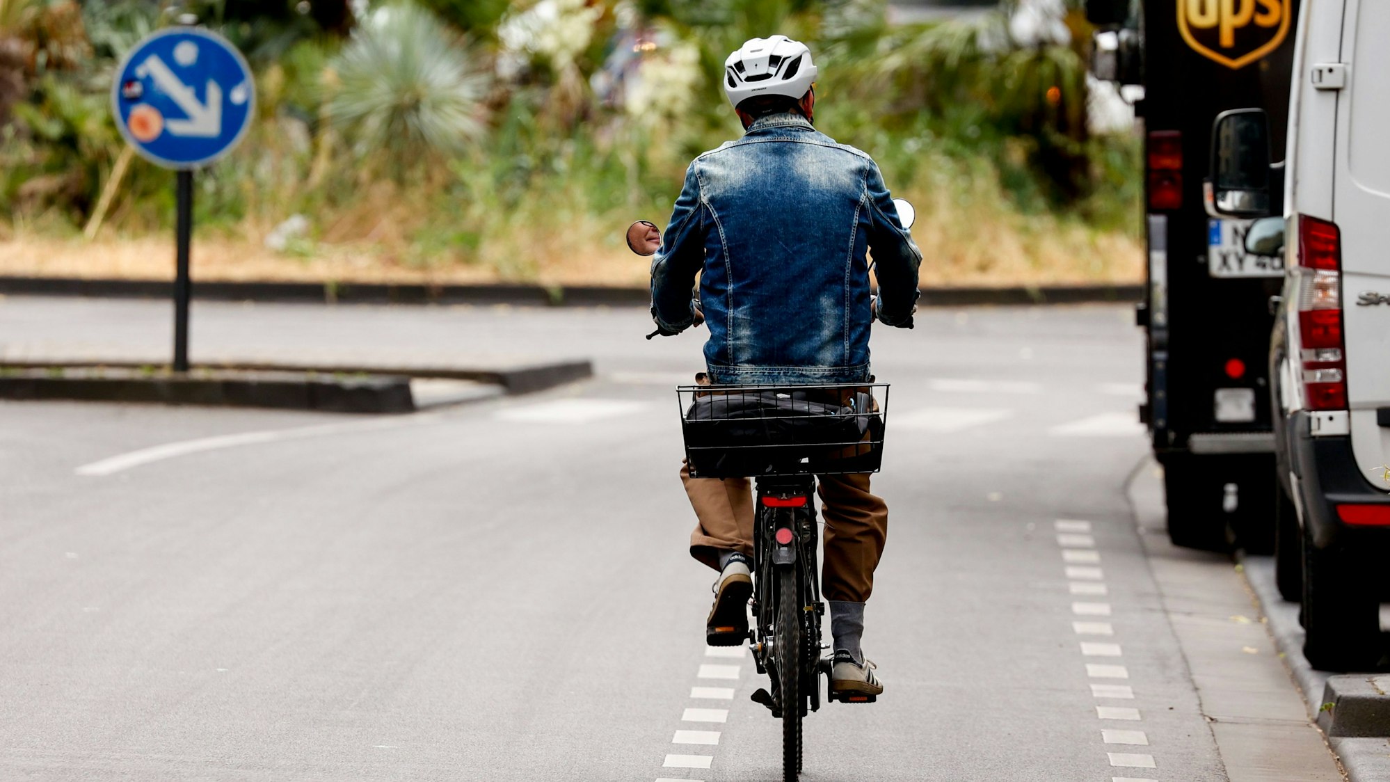 Zu sehen ist ein Radfahrer in der Rückansicht auf der Bonner Straße in Köln. Rechts daneben sind zwei Transporter geparkt.