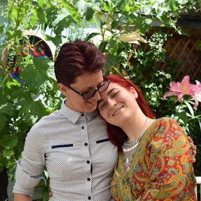 Adina Lupu (rechts) und Valeria Gavrilă stehen vor einem Hintergrund aus Pflanzen. Gavrilă lehnt an Lupus Schulter, lächelt in die Kamera. Lupu schaut sie an.