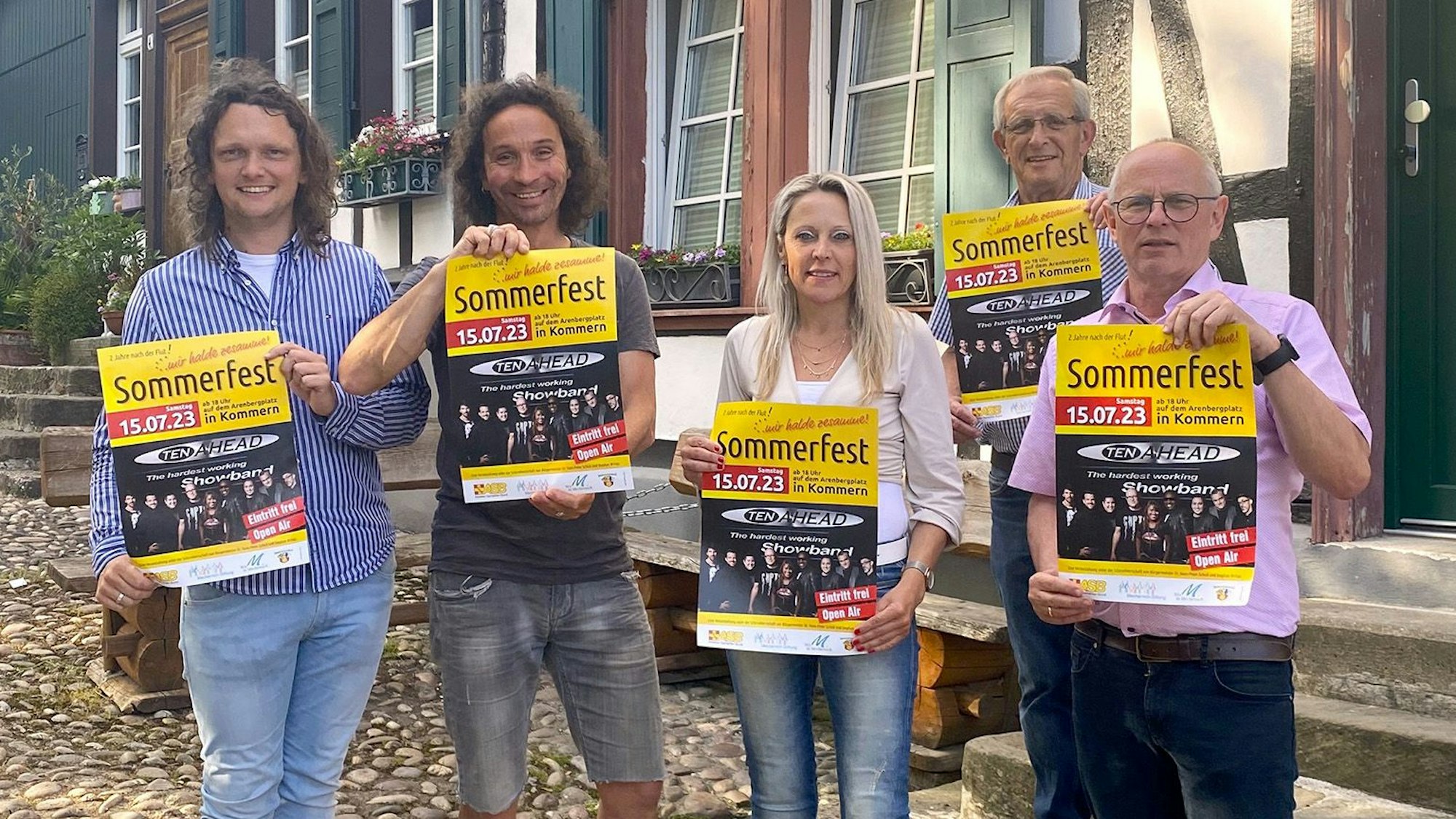 Mehrere Männer und eine Frau stehen vor einem Fachwerkhaus und zeigen Plakate einer Veranstaltung.