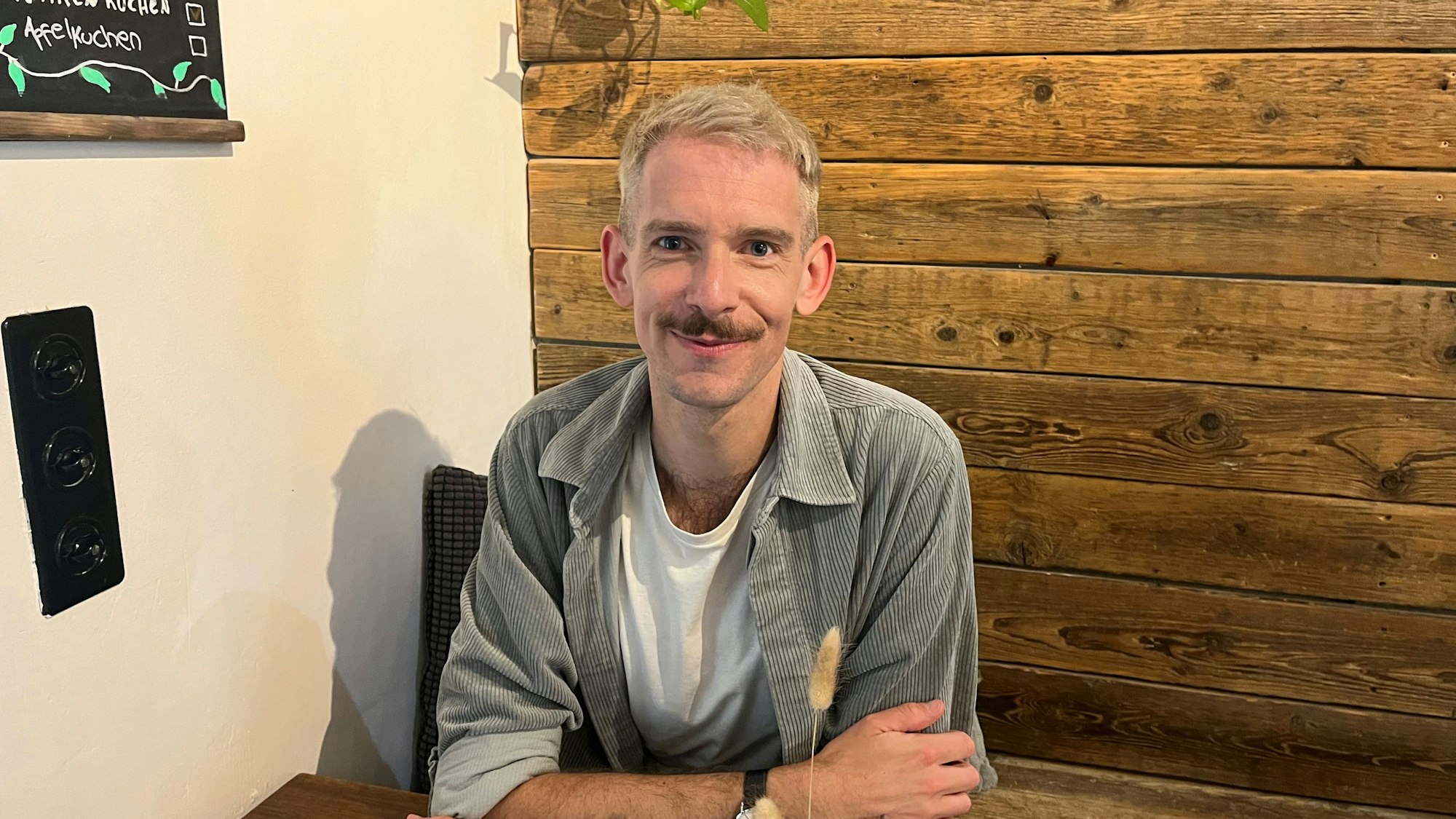 Ein junger Mann mit Schnurrbart sitzt an einem Tisch, mit den Armen über kreuz aufgestützt.