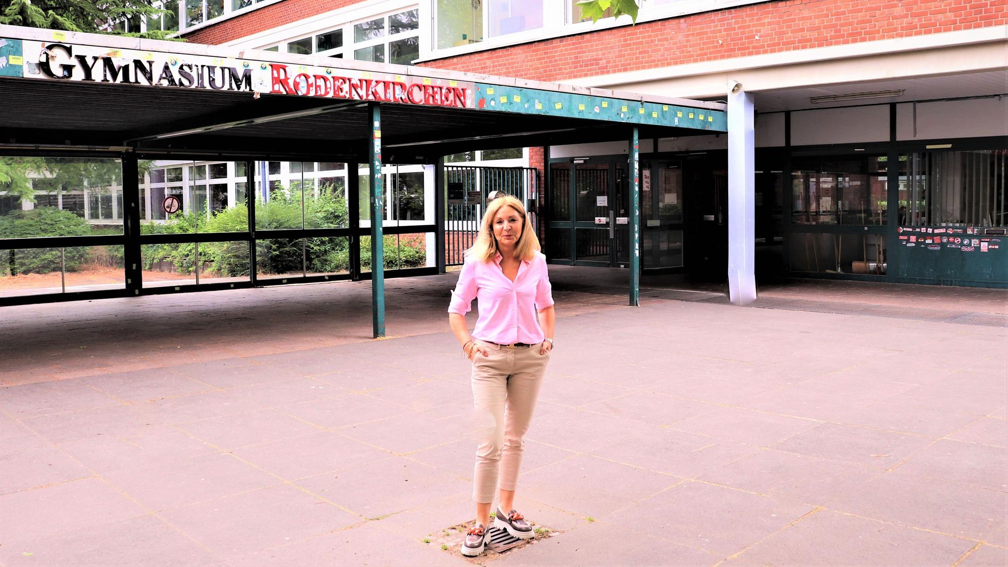 Eine blonde Frau mit schulterlangem blonden Haar steht vor einem Gebäude, auf dem der Schriftzug Gymnasium Rodenkirchen zu lesen ist.