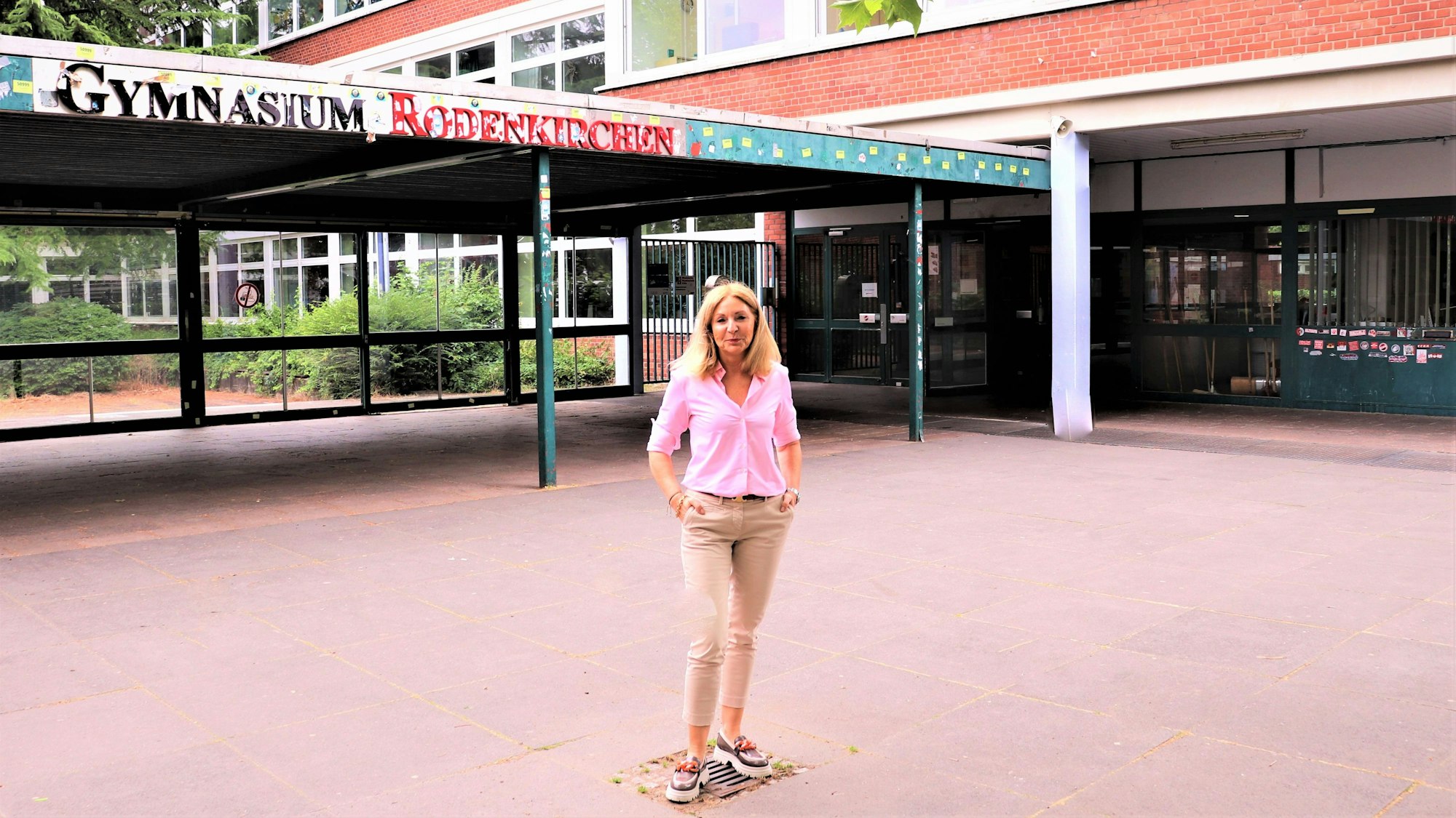Eine blonde Frau mit schulterlangem blonden Haar steht vor einem Gebäude, auf dem der Schriftzug Gymnasium Rodenkirchen zu lesen ist.