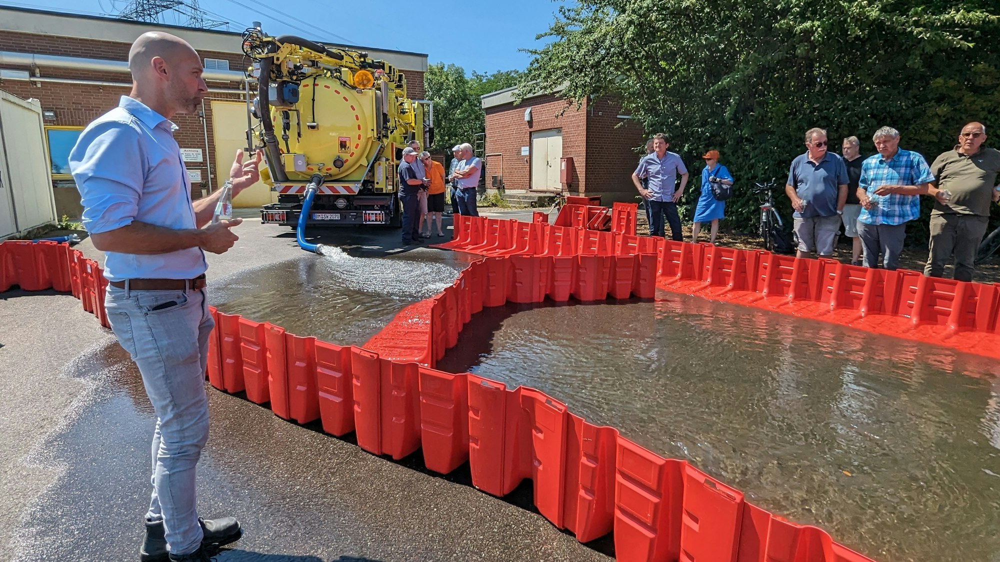 Ein Mann steht vor einer Reihe von zusammengesteckten Kunststoffelementen, die Wasser zurückhalten. Dahinter eine Menschengruppe.
