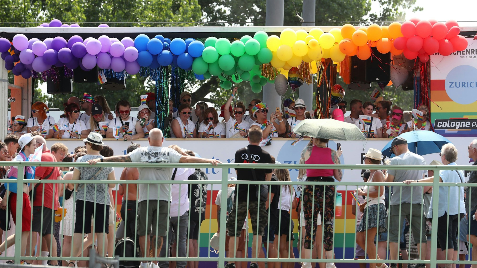 Ein Bus, der mit Luftballons in Regenbogenfarben geschmückt ist, fährt an Zuschauenden vorbei.