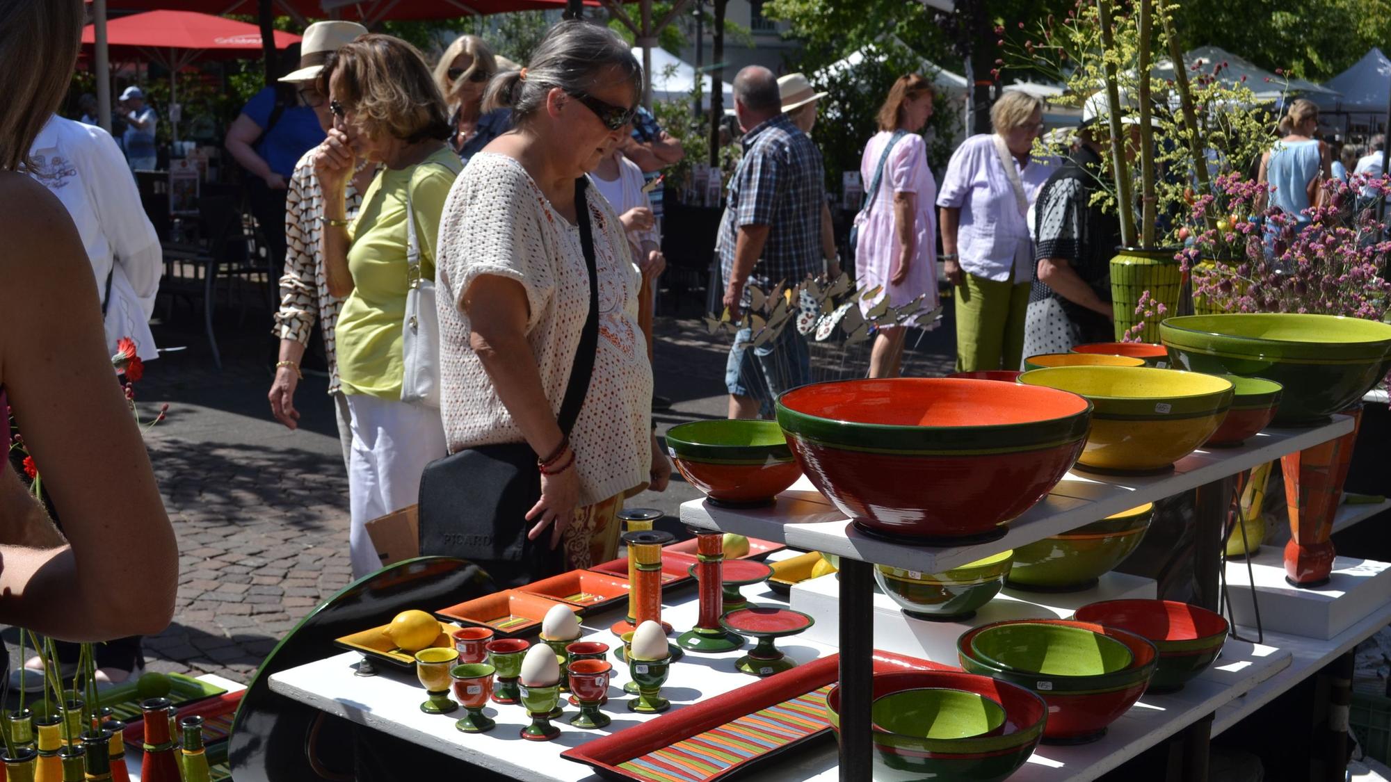 Bunte Vasen, Schüsseln und Eierbecher stehen auf einem mehrstöckigen Ausstellungstisch, an dem Besucher im Sonnenschein stehen.