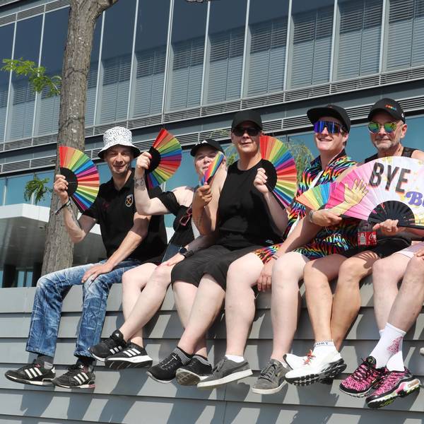 Sieben Teilnehmerinnen und Teilnehmer des Kölner CSD sitzen vor der Mauer eines Gebäudes und halten Fächer in Regenbogenfarben in der Hand.