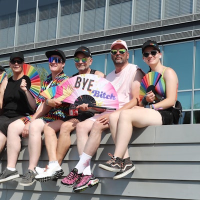Sieben Teilnehmerinnen und Teilnehmer des Kölner CSD sitzen vor der Mauer eines Gebäudes und halten Fächer in Regenbogenfarben in der Hand.