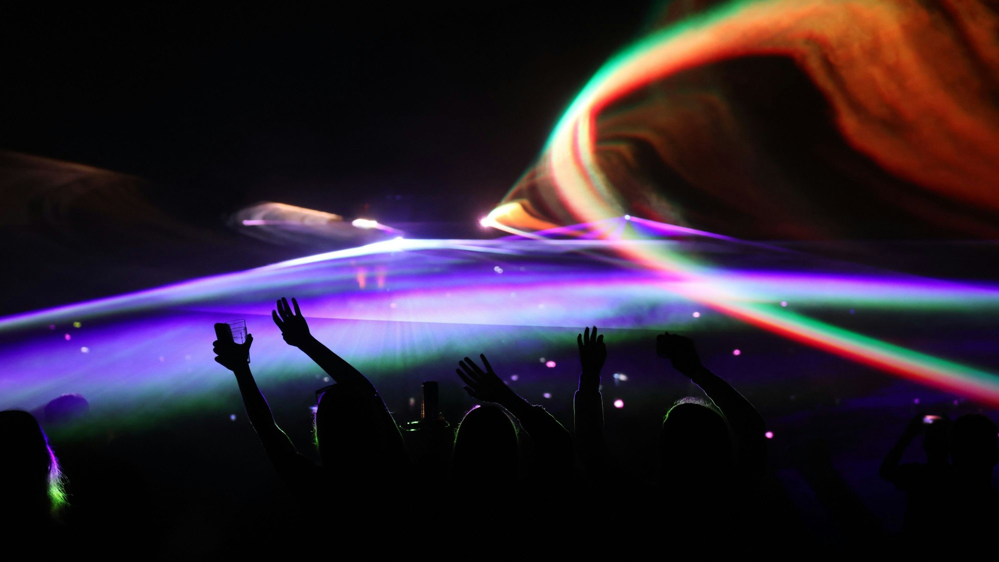 Menschen recken ihre Arme in die Höhe, farbige Lichter sind vor dunklem Nachthimmel zu sehen.