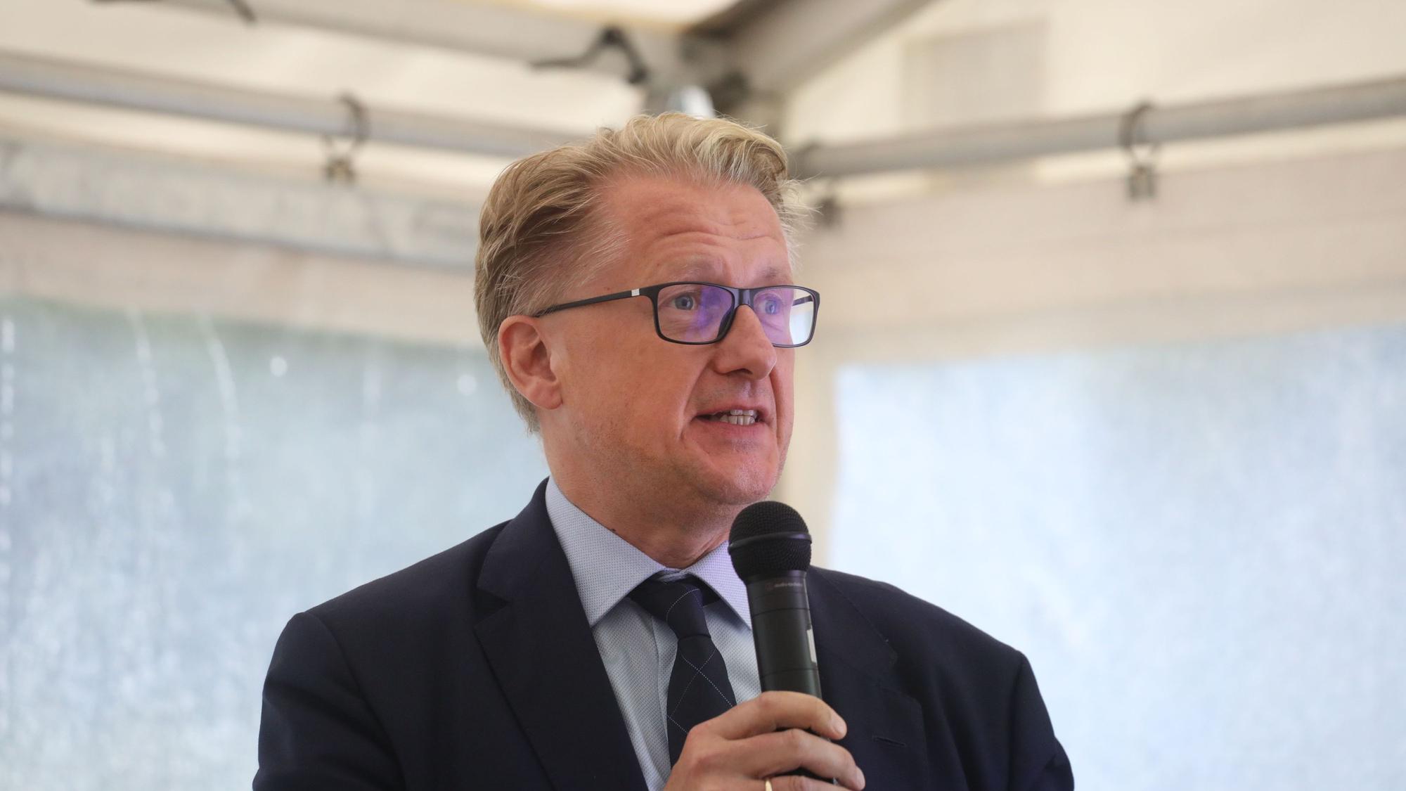 Ein Mann mit blonden Haaren und Brille hat ein Mikrofon in der Hand.