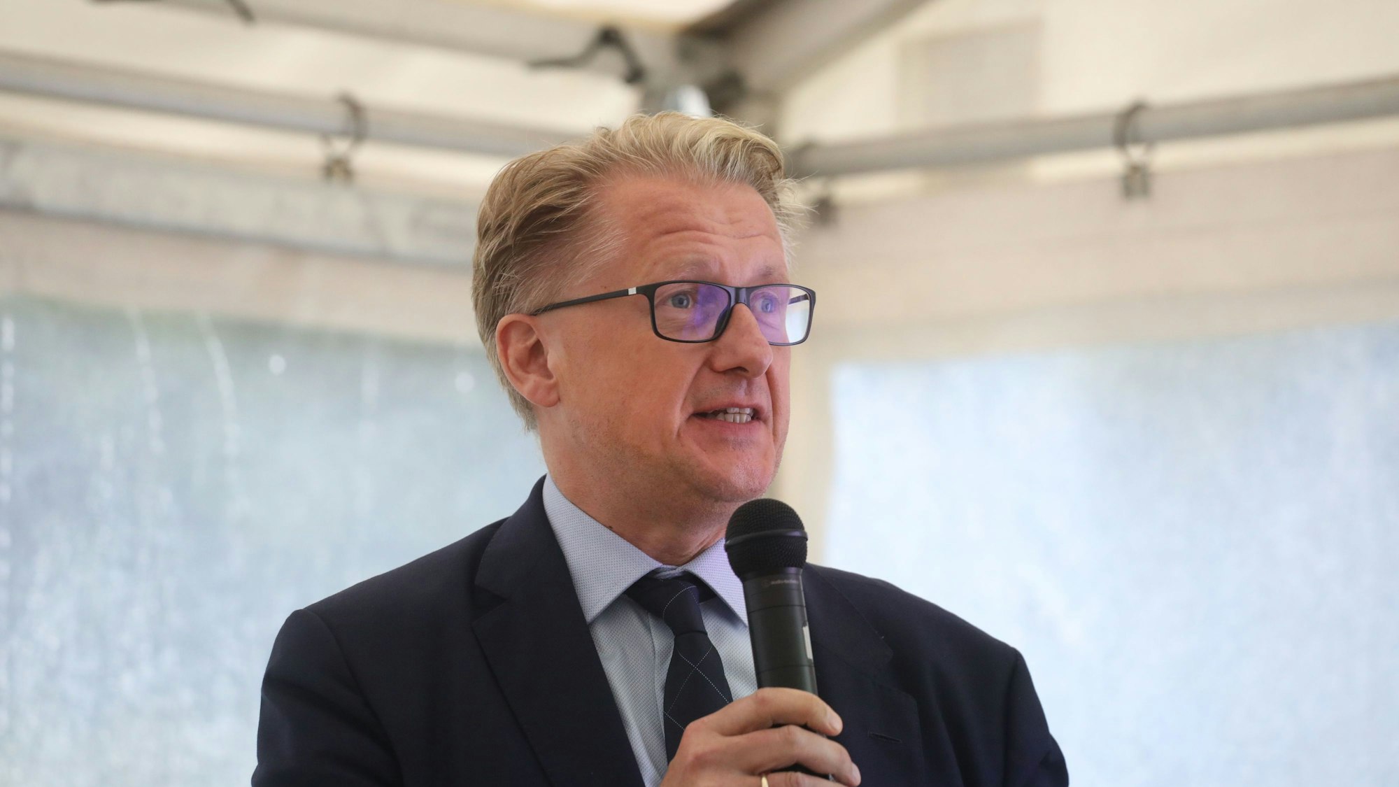 Ein Mann mit blonden Haaren und Brille hat ein Mikrofon in der Hand.