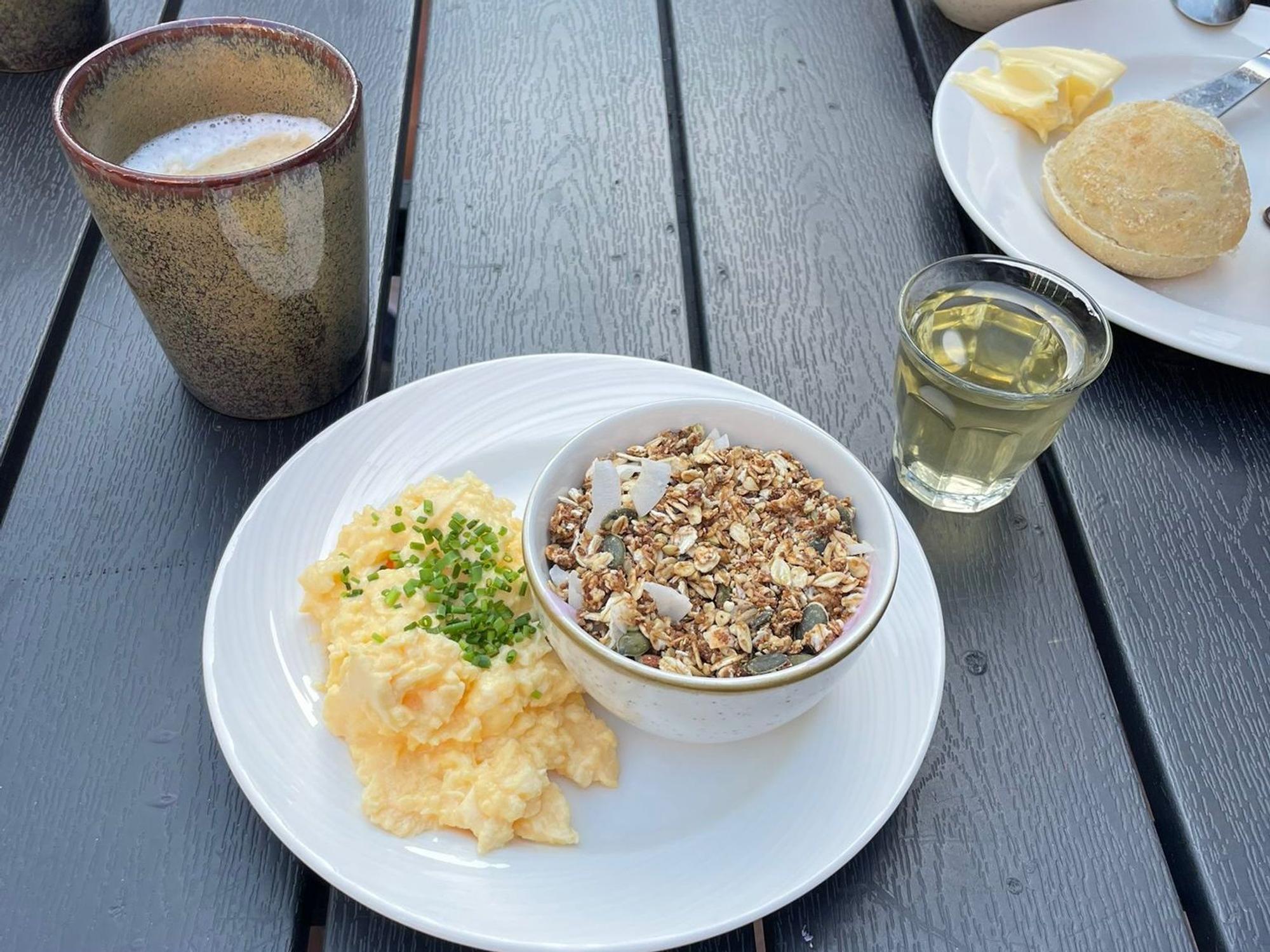 Frühstück: Auf einem Teller stehen eine gefüllte Müsli-Schale und Rührei, dahinter stehen ein Caffe Latte und ein Glas Apfelsaft. Im Hintergrund ein Teller mit einem Brötchen.