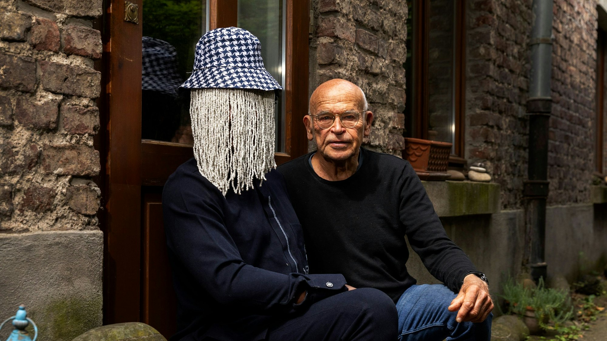 Die beiden sitzen nebeinander vor einer Tür. Anas trägt einen Hut mit einem Perlenvorhang, der sein Gesicht verdeckt.