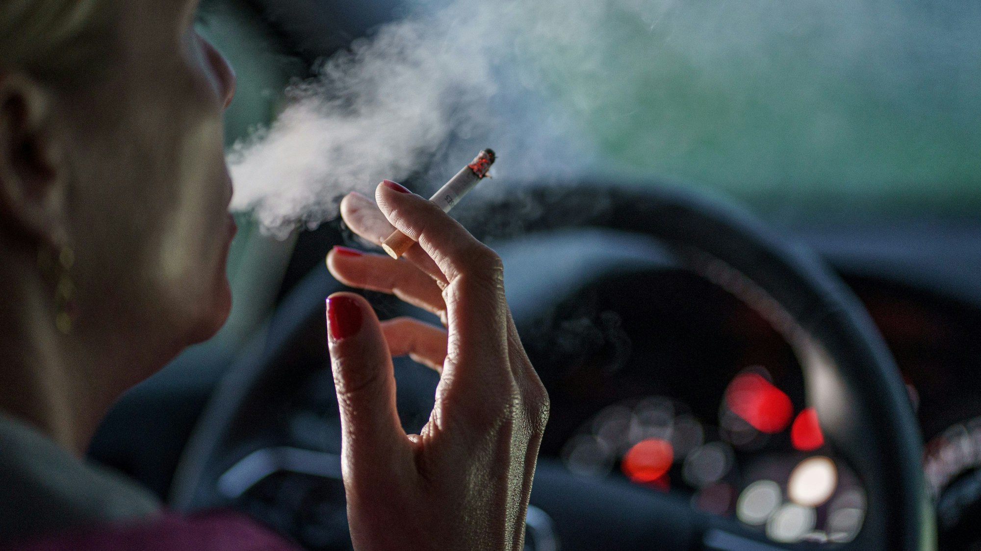 Eine Frau sitzt am Steuer eines Autos, hält in der rechten Hand eine Zigarette und bläst Rauch aus.