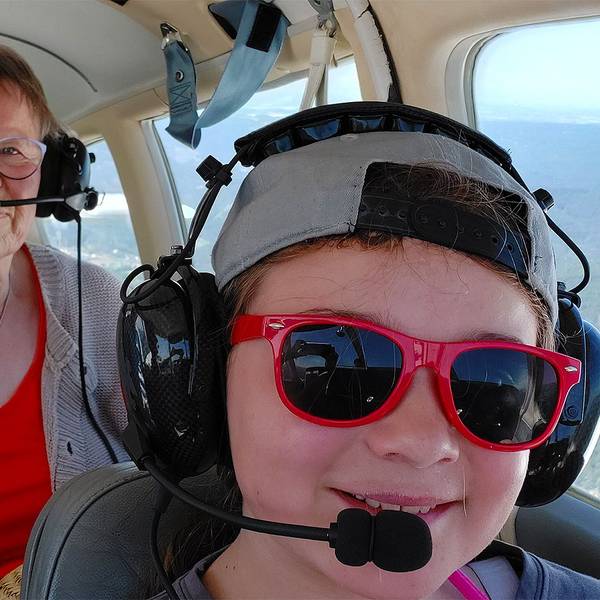 Das Bild zeigt ein Kind mit einer roten Sonnenbrille und eine Frau in einem kleinen Flugzeug. Beide tragen Kopfhörer mit Mikrofon.