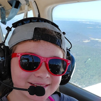 Das Bild zeigt ein Kind mit einer roten Sonnenbrille und eine Frau in einem kleinen Flugzeug. Beide tragen Kopfhörer mit Mikrofon.