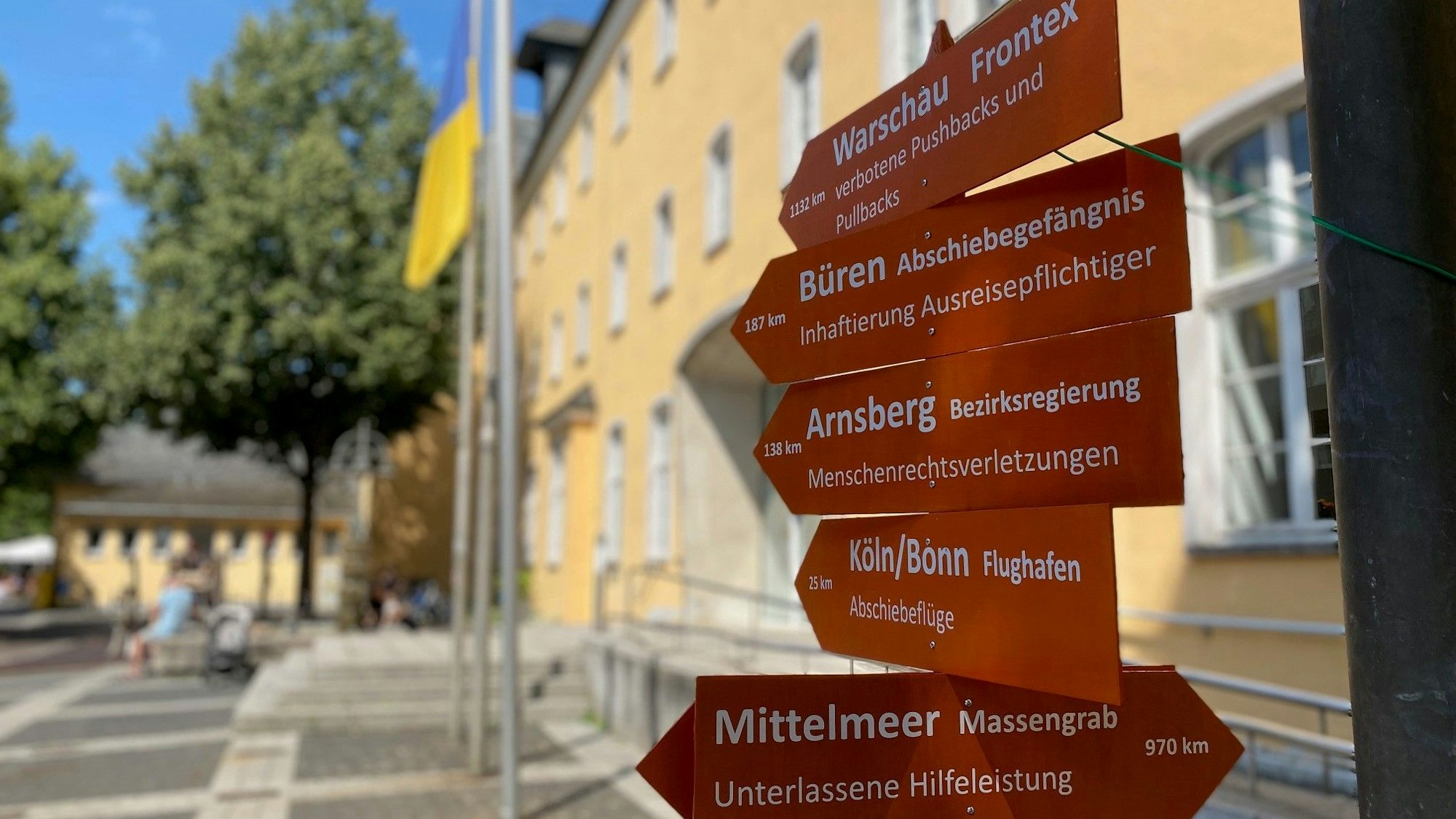 Die hölzernen Wegweiser tragen die Inschrift Mittelmeer, Massengrab und Flughafen Köln/Bonn Abschiebeflüge. Sie stehen vor dem Rathaus.