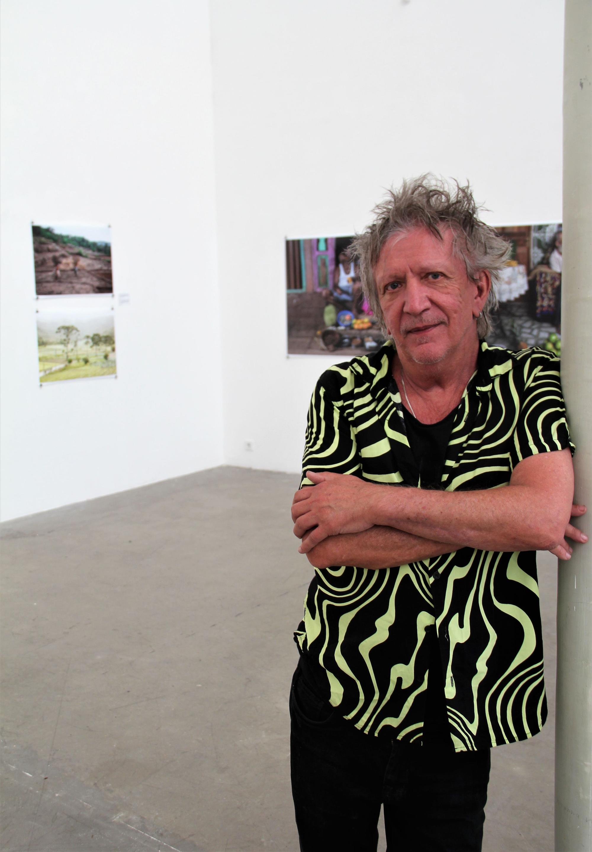Ein Man mit verstrubbelten grauen Haaren steht mit verschränkten Armen in einem Ausstellungsraum, in dem seine Bilder hängen.