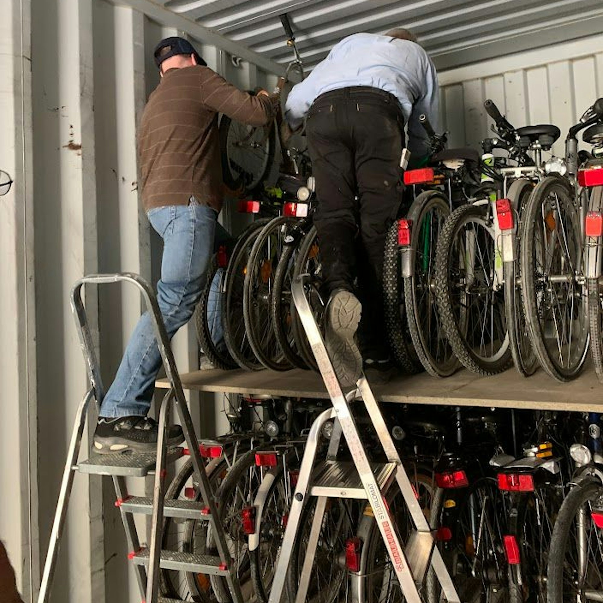 Zwei Männer stehen auf Leitern und stapeln Fahrräder in einem Container übereinander.