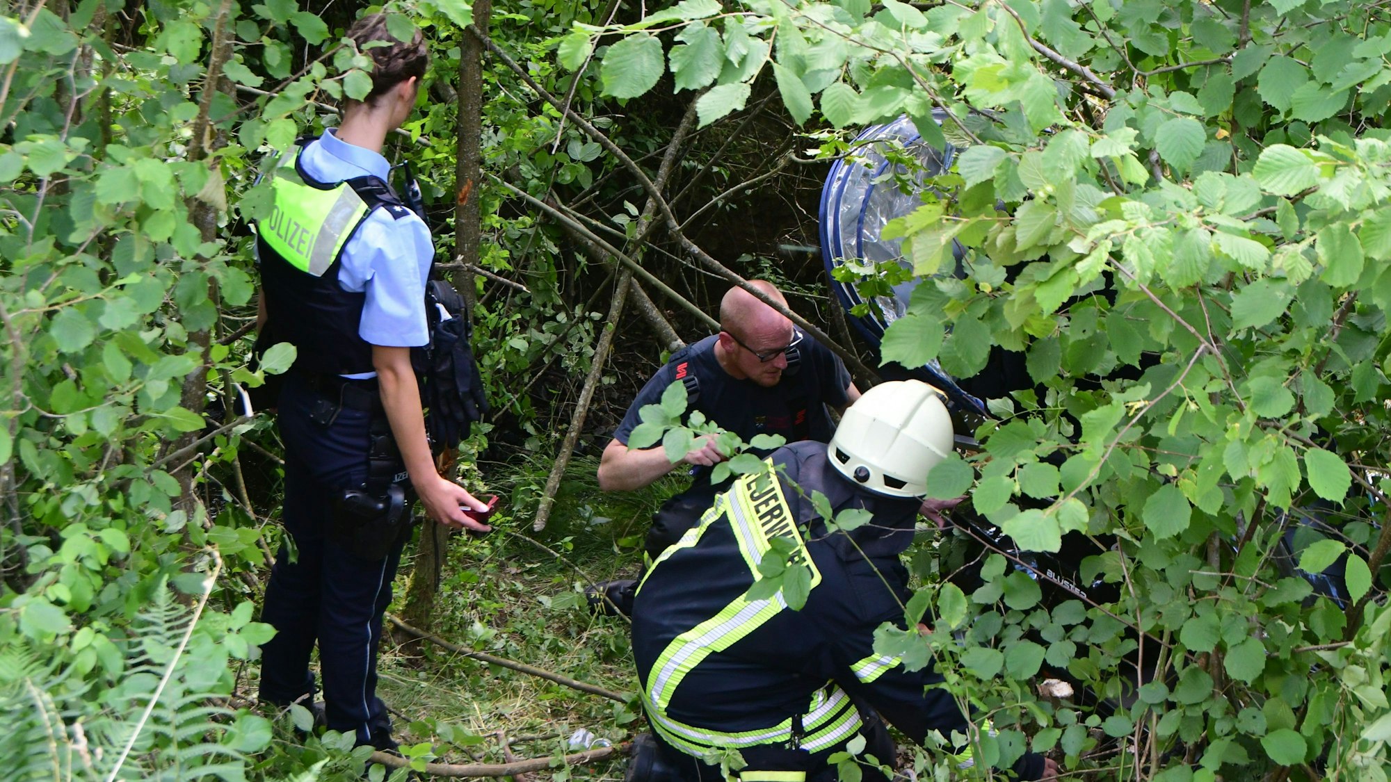 Das Bild zeigt eine Polizistin und zwei Feuerwehrleute, die eine Unfallstelle in dichtem Gestrüpp untersuchen.