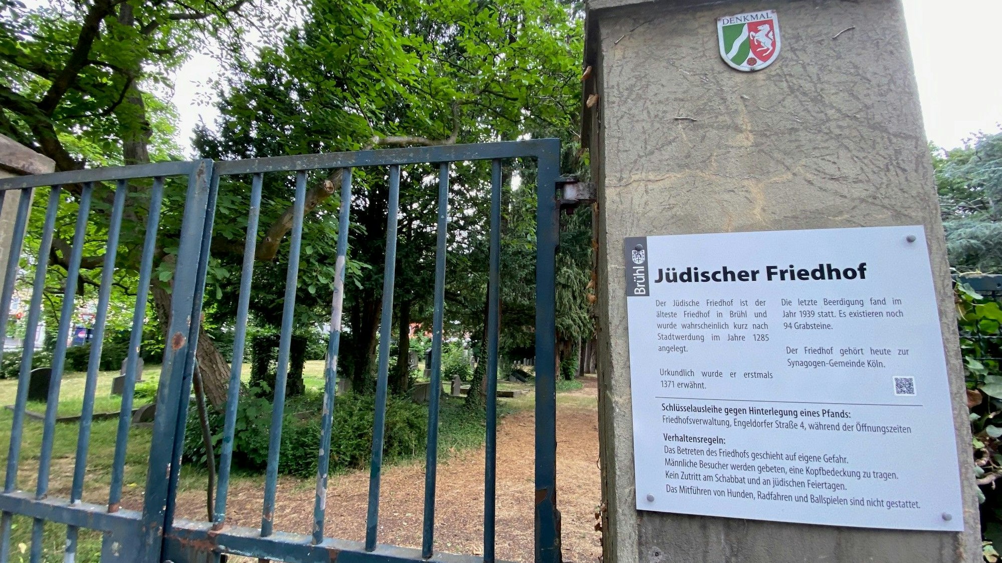 Das Tor am jüdischen Friedhof ist geschlossen, im Hintergrund sind Bäume und Grabsteine zu sehen.