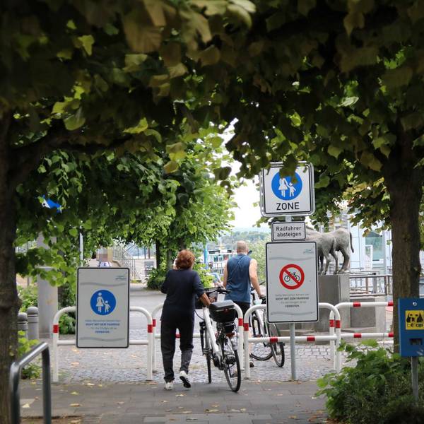 Zwei Radfahrer schieben ihre Räder auf der Rheinpromenade. Schilder weisen den Abschnitt in Höhe der Fähre als reine Fußgängerzone aus-