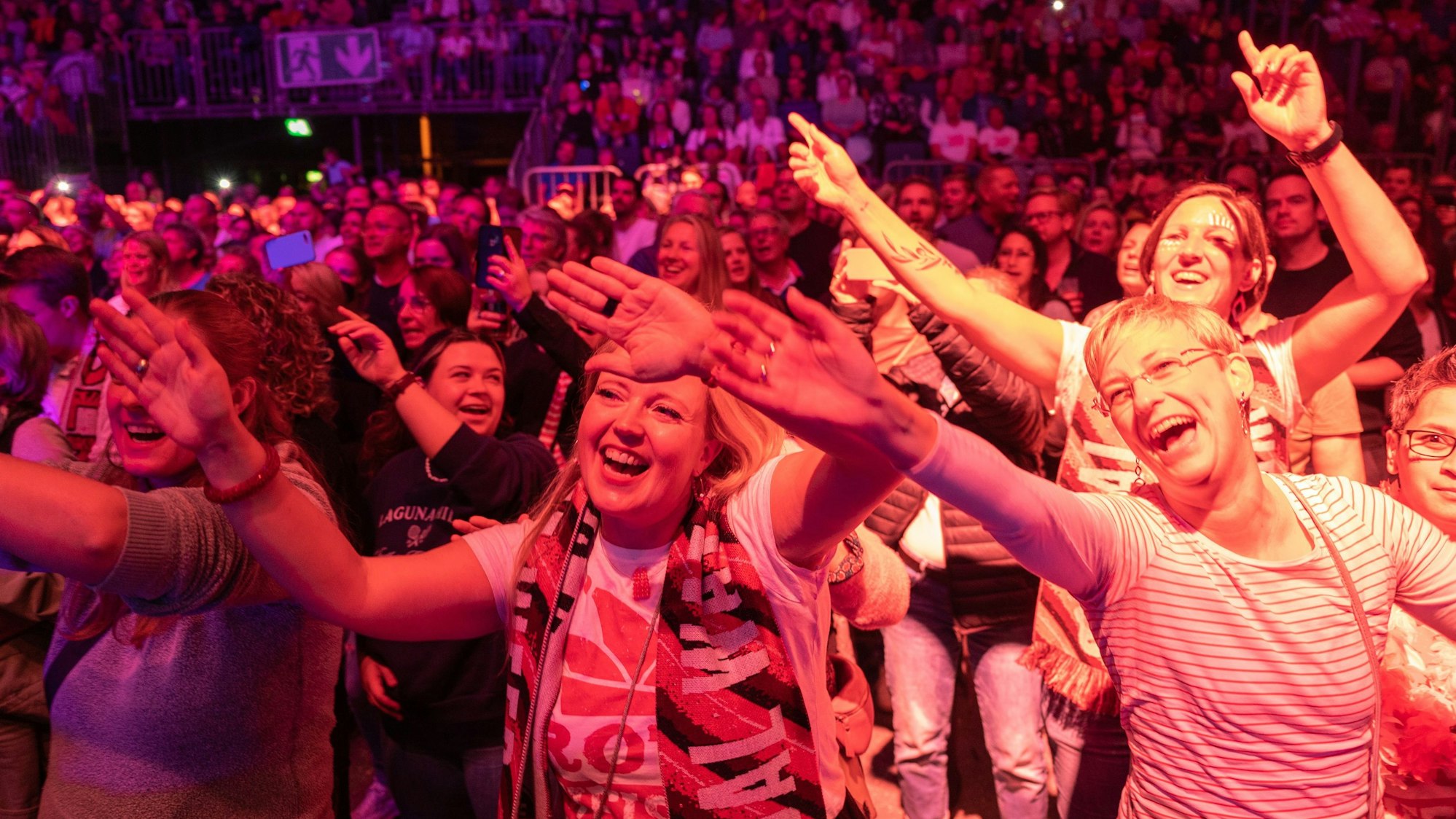 Im Vordergrund stehen schunkelnde, singende Frauen, die ihre arme in die Luft heben, im Hintergrund eine gefüllte Lanxess-Arena.