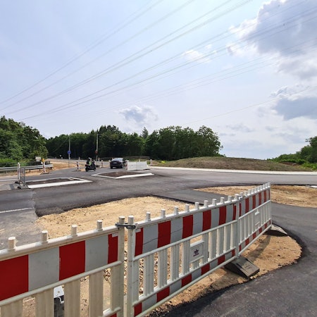 Das Bild zeigt eine frisch asphaltierte Straße, die oberste Asphaltschicht fehlt noch. Im Vordergrund stehen rot-weiße Warnbaken.
