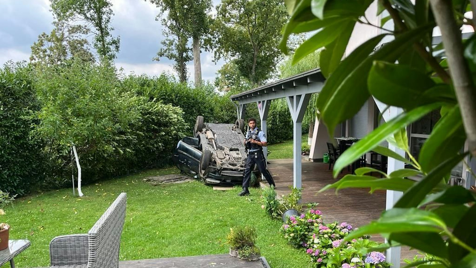 Ein Auto liegt in einem Garten auf dem Dach. Ein Polizist läuft umher.