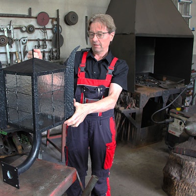 Das Bild zeigt Schmied Albert Köller in seiner Werkstatt. Er hat eine große Lampe in der Hand.