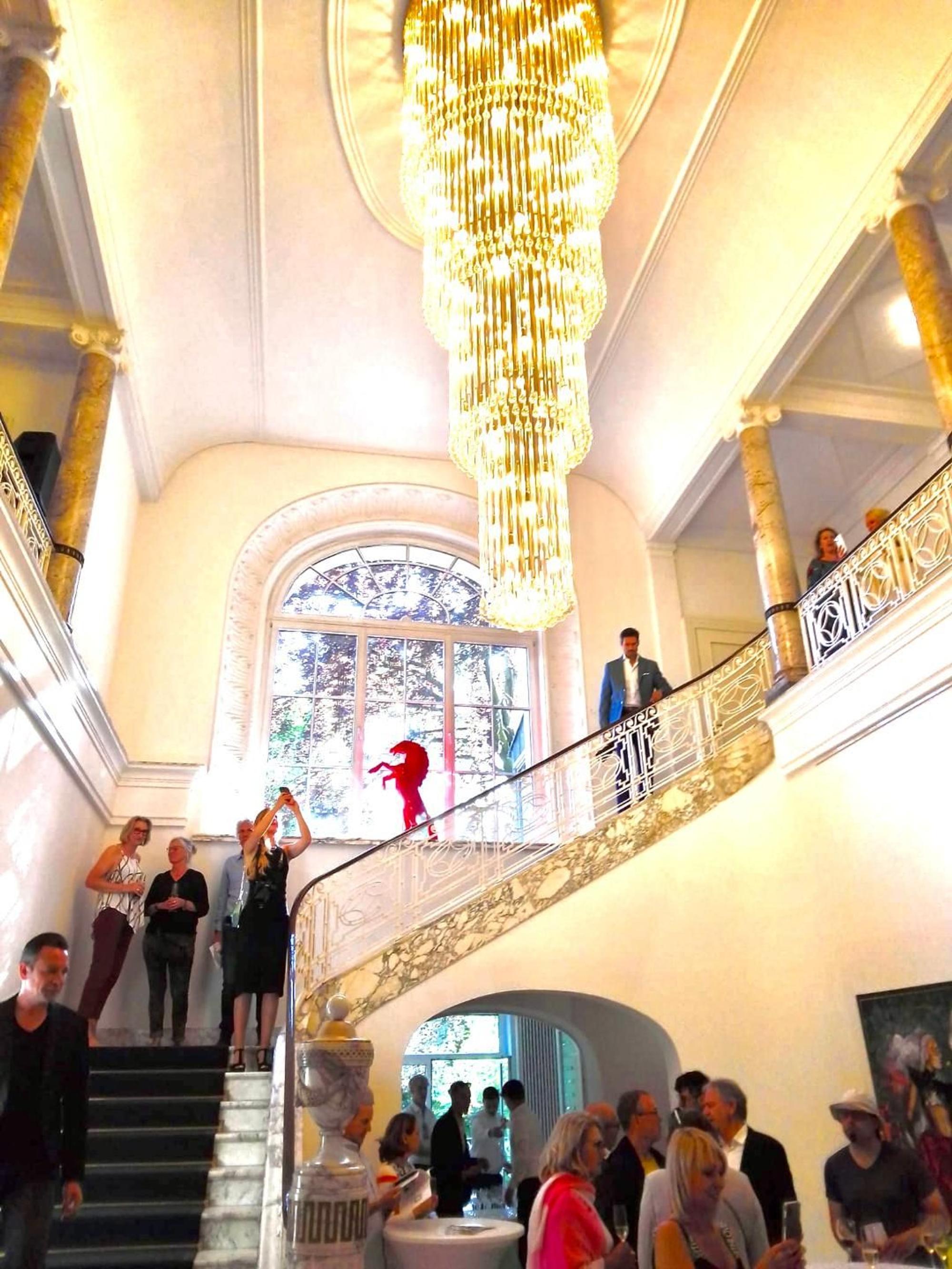 Ein großzügiges Treppenhaus ist zu sehen, Menschen mit Gläsern stehen im Raum unten und auf der Treppe. Bilder hängen an der Wand, Skulpturen sind aufgestellt.
