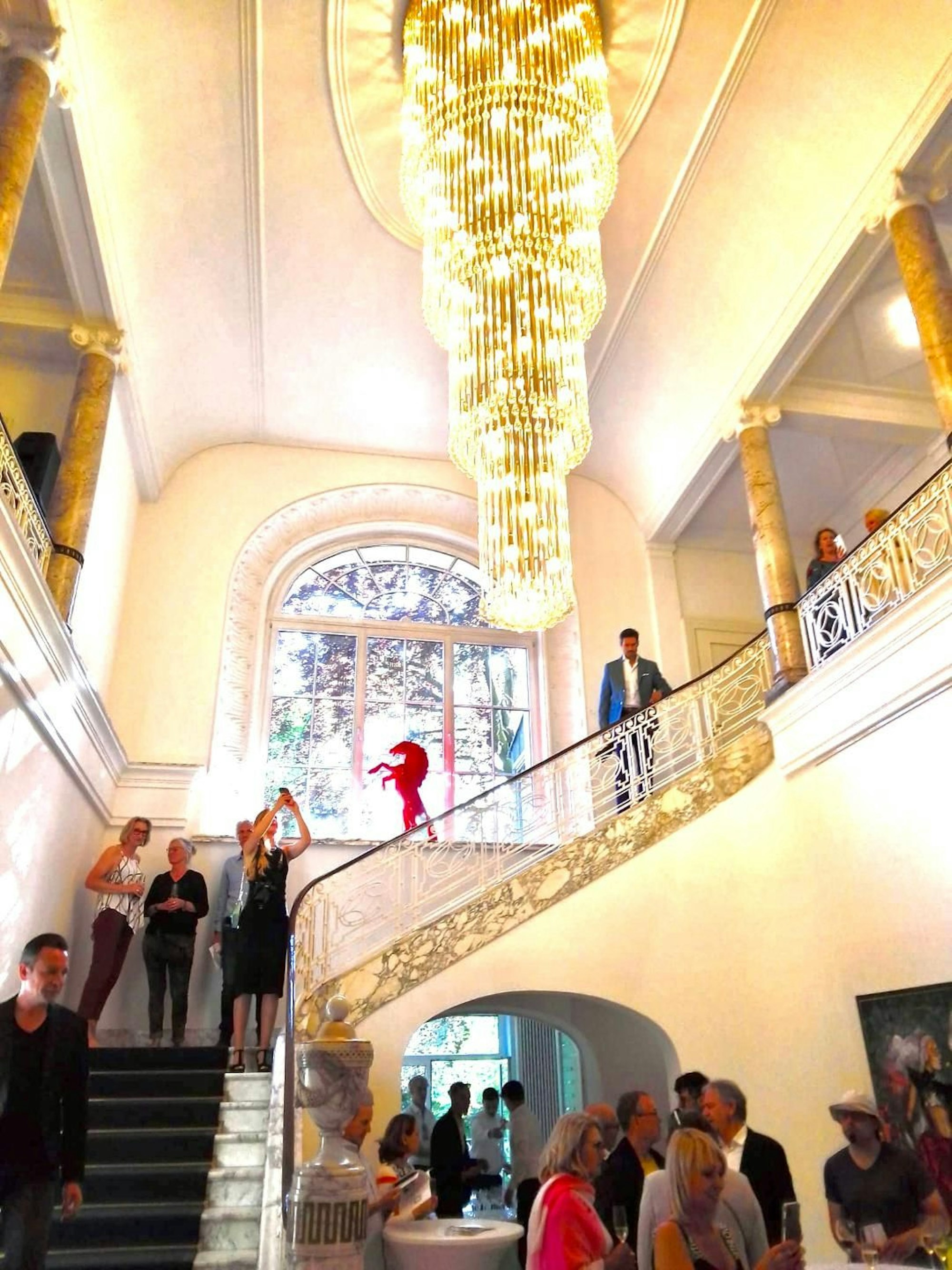 Ein großzügiges Treppenhaus ist zu sehen, Menschen mit Gläsern stehen im Raum unten und auf der Treppe. Bilder hängen an der Wand, Skulpturen sind aufgestellt.