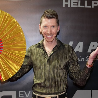 Manuel Flickinger auf der All Together Charity-Gala 2023. Er hält einen großen goldenen Fächer in der Hand.