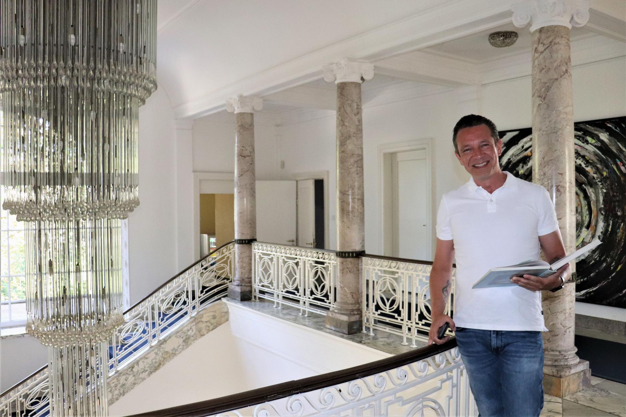 Ein Mann im weißen Polohemd und Jeans steht im marmornen Treppenhaus.