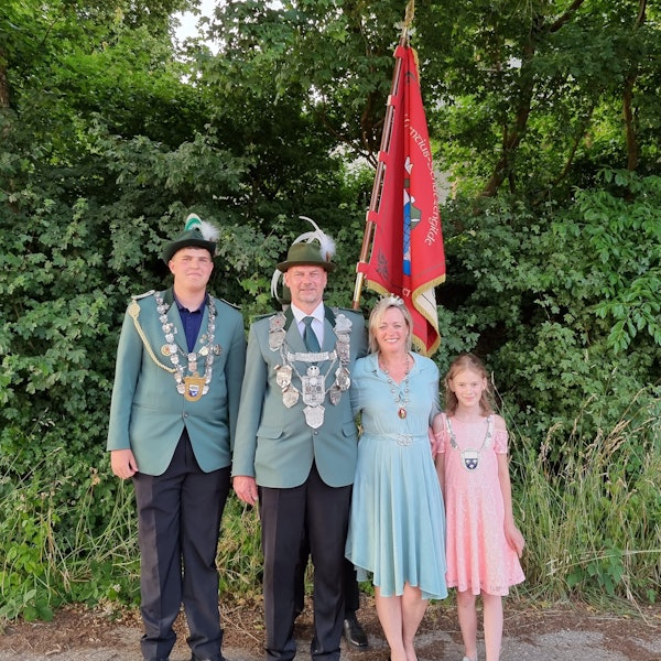 Zwei Männer, eine Frau und ein Mädchen stehen vor einer Hecke. Die Männer tragen Schützenuniform. Hinter den vier Menschen steht eine rote Fahne.