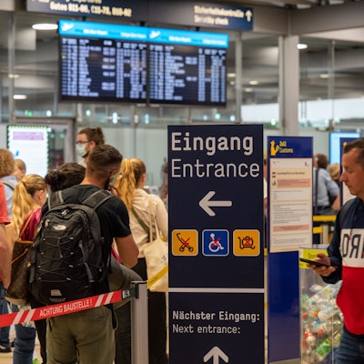 Am Flughafen Köln/Bonn warten Reisende im Sommer 2022 an der Sicherheitskontrolle. Es haben sich extrem lange Schlangen gebildet, im Hintergrund ist ein Fluganzeiger zu sehen. (Archivbild)