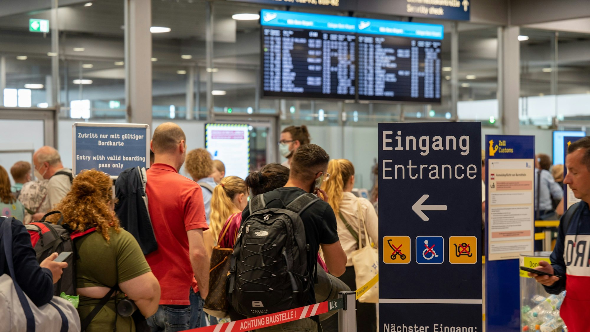 Am Flughafen Köln/Bonn warten Reisende im Sommer 2022 an der Sicherheitskontrolle. Es haben sich extrem lange Schlangen gebildet, im Hintergrund ist ein Fluganzeiger zu sehen. (Archivbild)
